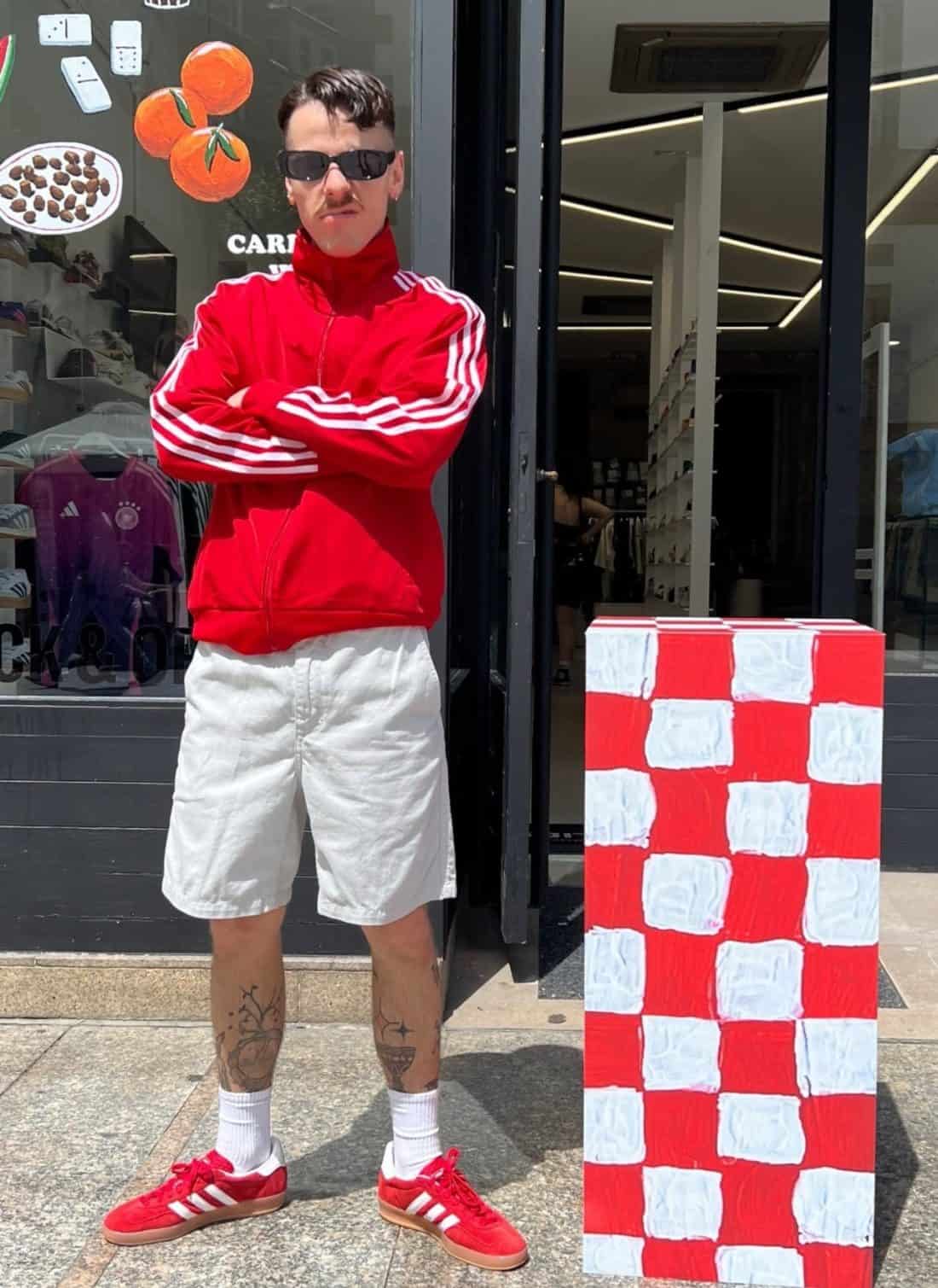 Ataviada con el atrevido conjunto Festival Fever (una chaqueta deportiva roja vibrante combinada con pantalones cortos blancos y zapatillas rojas a juego), una persona se para con seguridad, con los brazos cruzados, y exuda una innegable sensación de fiebre festivalera. Luciendo gafas de sol y exhibiendo tatuajes en las piernas, se encuentra de pie junto a una llamativa escultura a cuadros rojos y blancos.