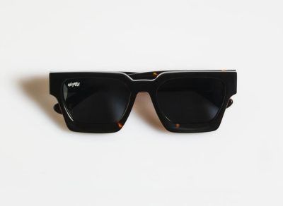 Gentleman Guy, un par de elegantes anteojos de sol cuadrados negros con lentes oscuros, se presenta sobre un fondo blanco liso. Perfectos para el caballero moderno, estos marcos gruesos y brillantes ofrecen una apariencia elegante que complementa cualquier atuendo.
