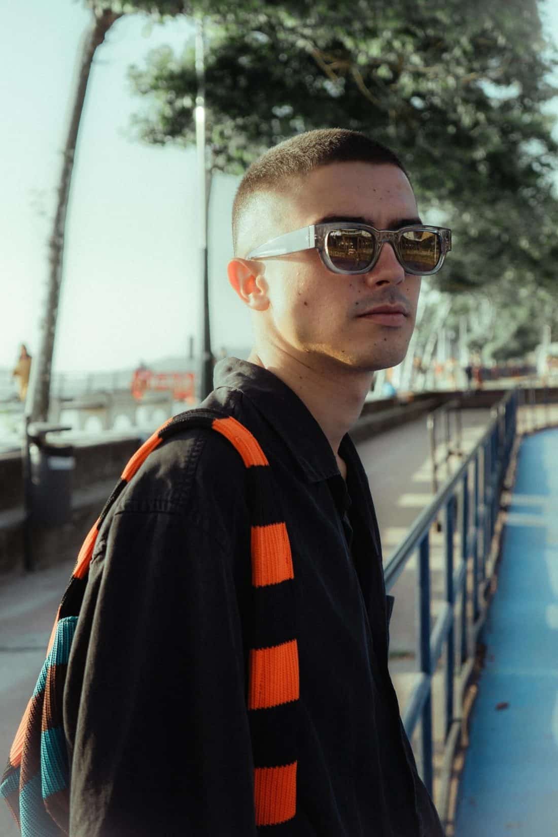 Una persona con el pelo corto, gafas de sol y una camiseta negra, está de pie junto a la costa y derrocha estilo. Lleva sobre el hombro la bufanda Radical Roots, con rayas naranjas y negras llamativas que reflejan la esencia del otoño. Al fondo, los árboles enmarcan la tranquila imagen borrosa del agua.