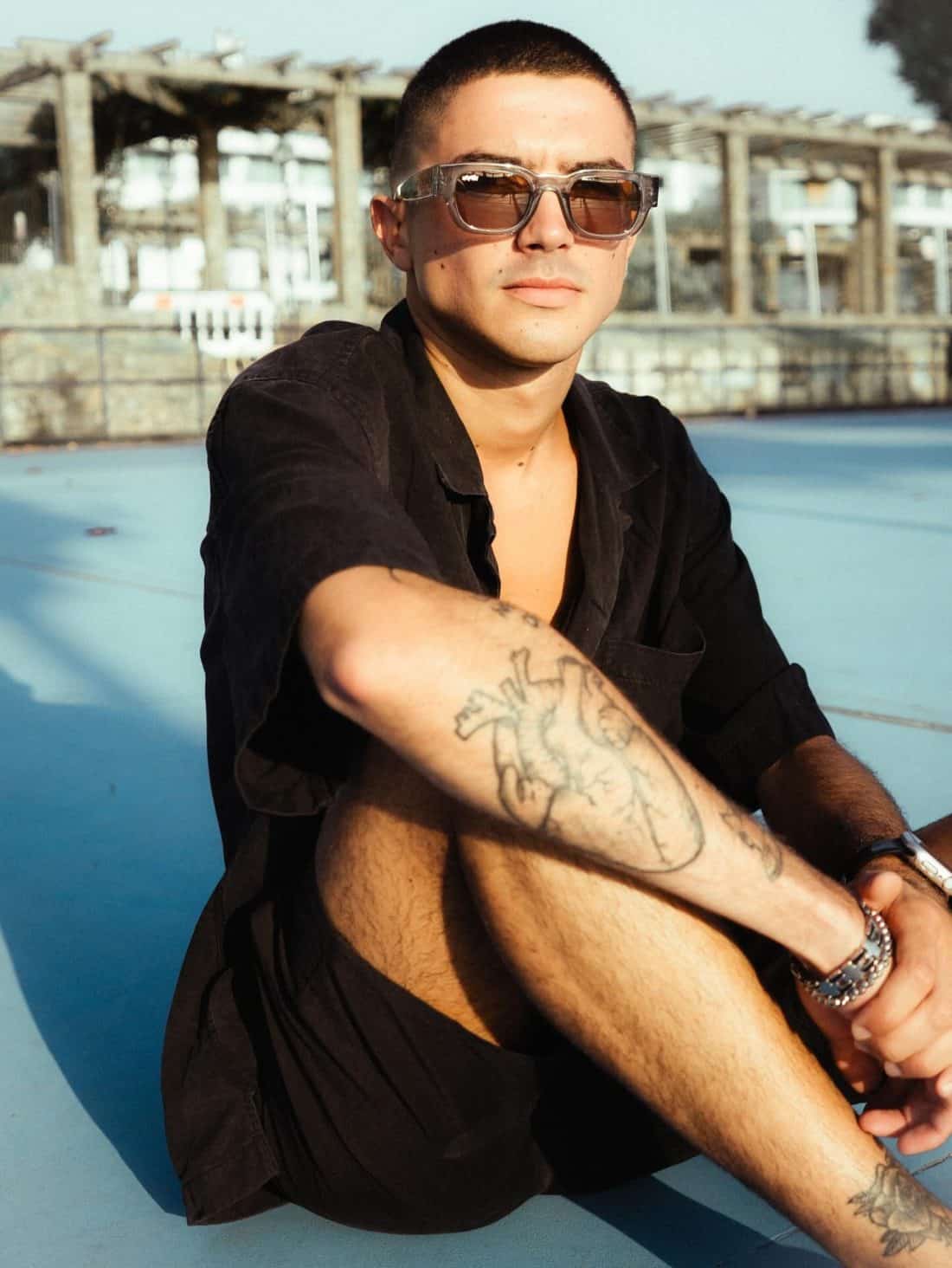 Una persona con cabello corto y gafas de sol se relaja en una superficie azul al aire libre, vistiendo una camiseta negra y pantalones cortos que resaltan sus tatuajes Radical Roots en el brazo y la pierna. Al fondo, una estructura con vigas de madera realza el ambiente radical.