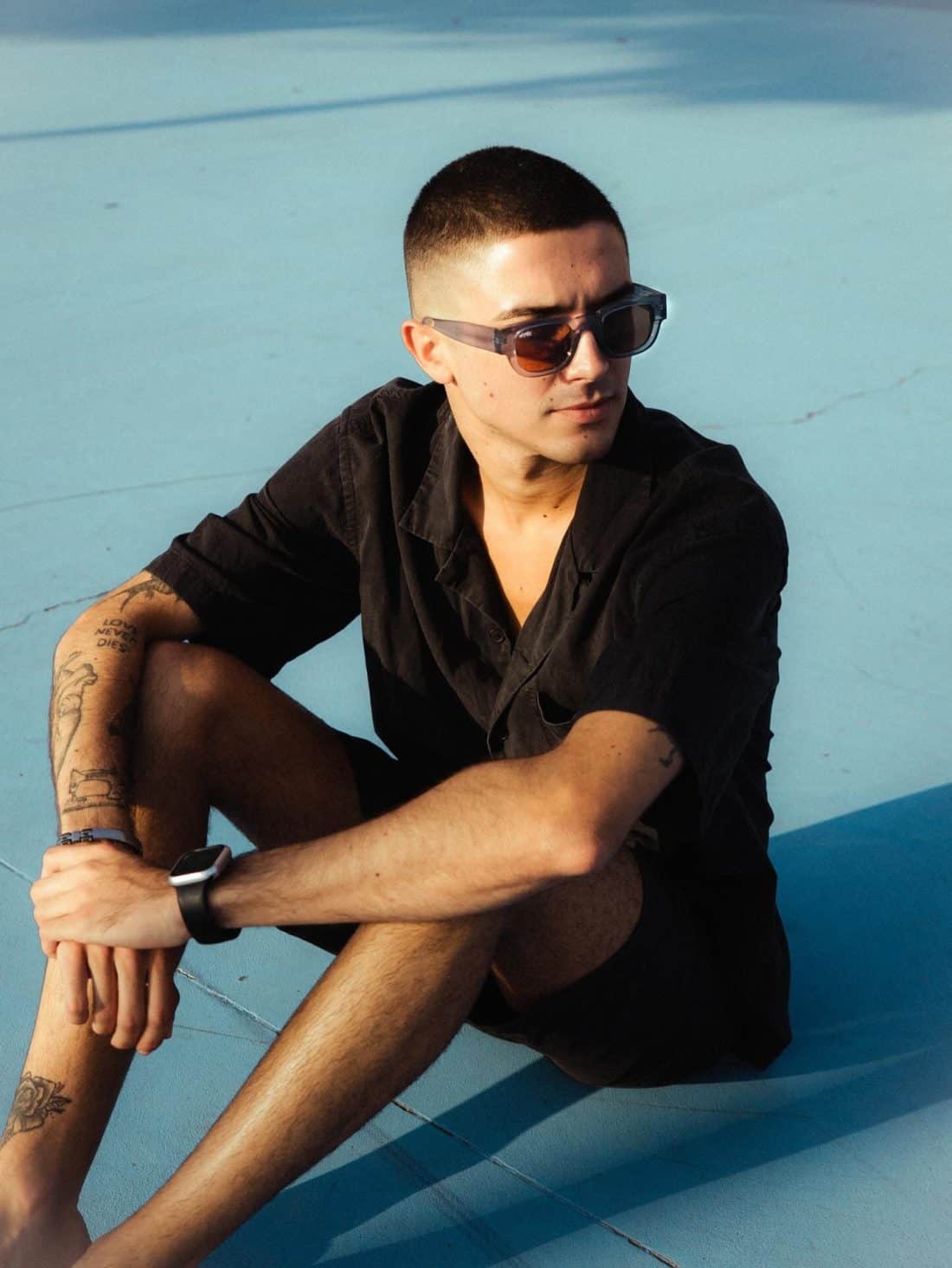 Una persona con cabello corto y gafas de sol se sienta sobre una superficie azul claro, luciendo el atuendo Radical Roots. Su camisa y pantalones cortos negros resaltan los tatuajes que trazan sus raíces a lo largo de sus brazos y piernas, exudando una vibra verdaderamente radical.
