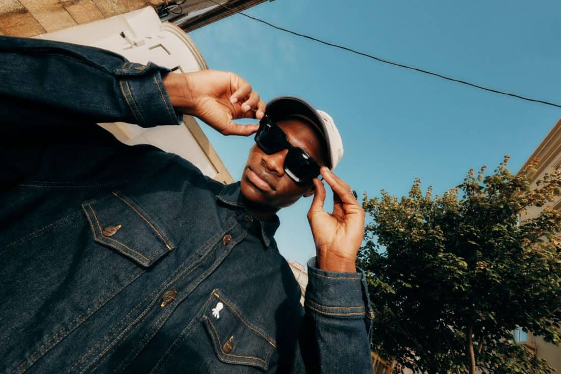 Un caballero conocido como "Gentleman Guy", con gafas de sol y gorra, se encuentra al aire libre frente al cielo azul claro. Lleva una chaqueta de mezclilla oscura y usa ambas manos para ajustar sus gafas de sol, con árboles y edificios que brindan un pintoresco telón de fondo.