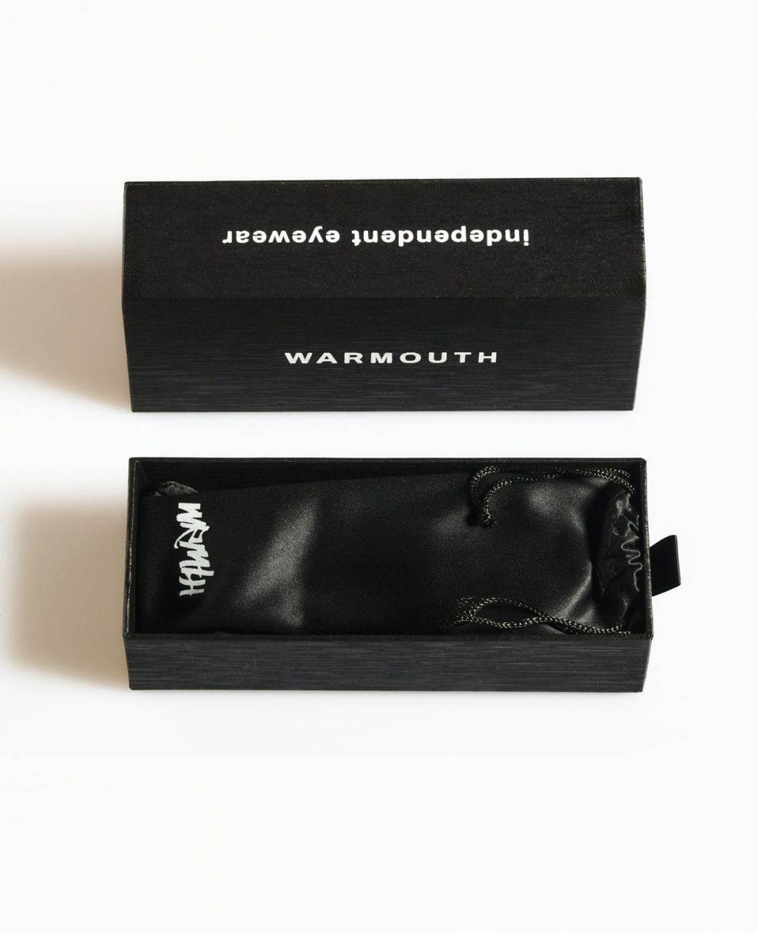 La caja negra, en la que se destaca el nombre "WARMOUTH", contiene la colección de gafas Radical Roots. En el interior, se ve parcialmente una bolsa negra con cordón adornada con un logotipo blanco. La caja abierta se presenta elegantemente sobre una superficie blanca prístina.
