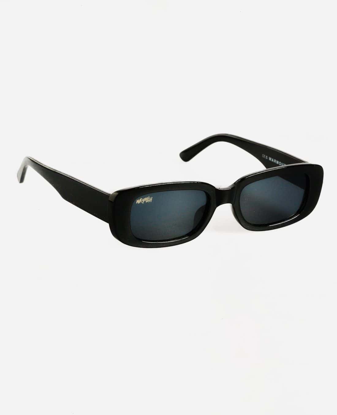 Un par de gafas de sol Festival Fever, con un diseño elegante y moderno con marcos rectangulares negros, lentes oscuras y el nombre de la marca "KOMONO" sutilmente impreso en ellas, son perfectas para la temporada de festivales. Están fotografiadas sobre un fondo blanco liso.