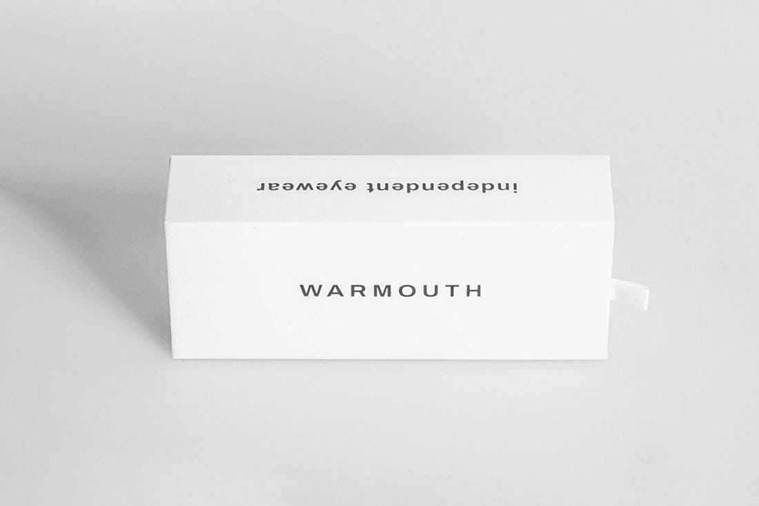 Una caja rectangular blanca que muestra de manera destacada la palabra "WARMOUTH" en el frente y la palabra "independent eyewear" en la parte superior, se muestra al revés mientras representa a Radical Roots sobre un fondo gris simple, proyectando una sombra suave hacia la izquierda.