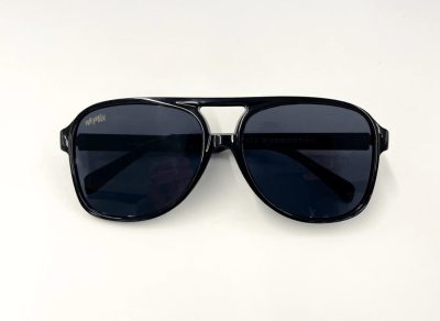 Las gafas de sol Cowboy Cool 2.0, con montura negra brillante estilo aviador y lentes oscuras, irradian un estilo robusto y elegante sobre un fondo blanco. Su elegante diseño se ve realzado por un distintivo puente doble, que le suma un encanto moderno.